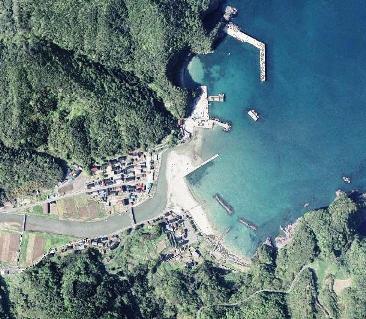 緑の山と青い海が広がり、山の麓の漁港近くに家々が並び、漁港からは堤防が伸びている浦島漁港の航空写真