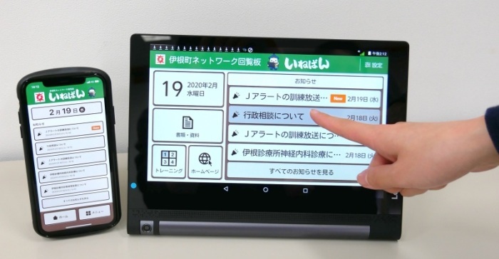 スマートフォンとタブレット端末の画面に伊根町ネットワーク回覧版「いねばん」のトップ画面が表示され、指でタブレット端末の画面を操作しようとしている写真