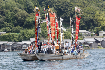 赤や黒などの綺麗な飾り旗が飾られた2艘の祭礼船に、頭に赤いハチマキをまき、祭りの衣装を身に着けた多くの人が乗っている祭りの様子写真