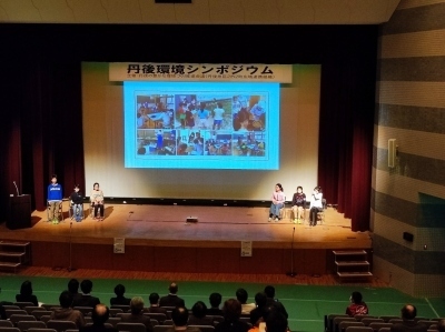 ステージ上の大きなスクリーンに写真が映し出され、スクリーンの右端と左端にそれぞれ3人の児童がステージ上に上がり発表をしている写真