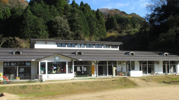 園舎の後方には緑豊かな木々が見え、白壁にこげ茶色の屋根で横長に造られた園舎と前方に広々としたした園庭が見える伊根保育園の外観写真