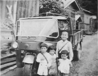 前輪が1つの三輪トラックの前に4人の子供たちが立っている昭和30年頃の白黒写真